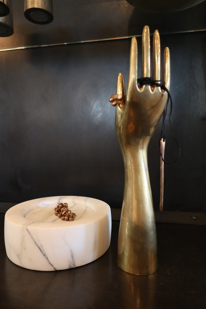 A brass hand-shaped stand holds a thin black string with a Pistilli 01 ring by Monica Castiglioni, while a marble dish displays another Monica Castiglioni ring featuring round details, all set against a dark backdrop.